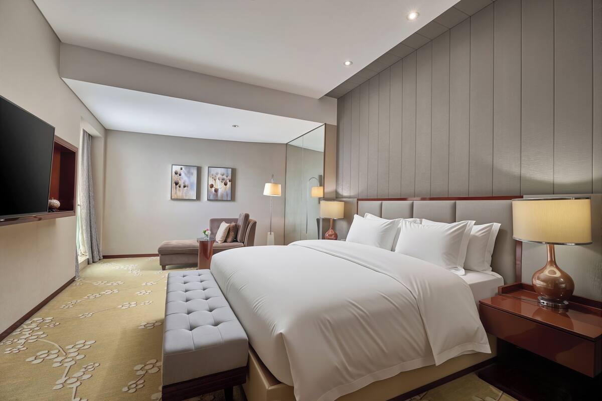 A modern hotel room with a large bed, a seating area, a television, and a mirror.