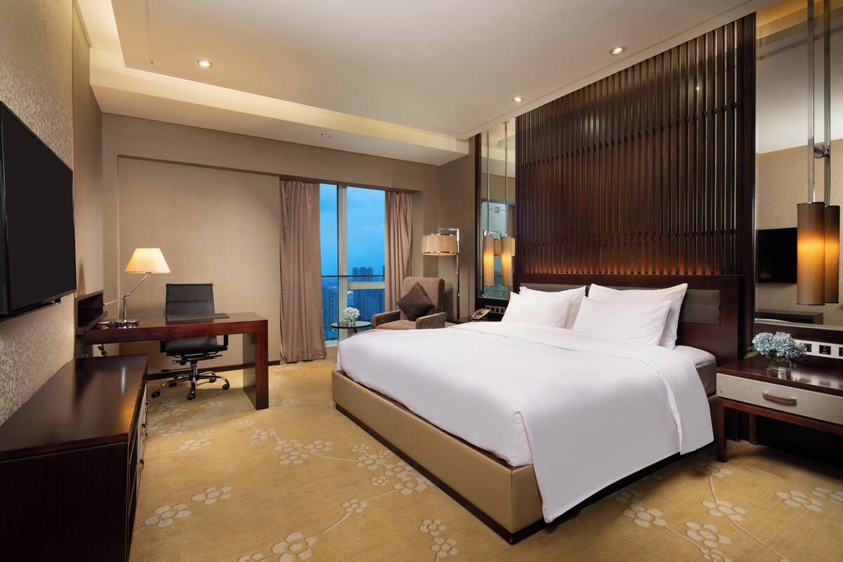 A hotel room with a large bed, a desk with a chair, a TV, and a view of the city.