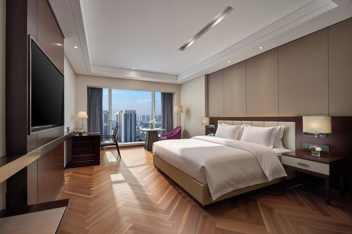 A modern hotel room with a large bed, a TV, a desk, and a view of the city.