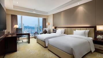 A hotel room with two beds, a desk, and a large window offering a city view.