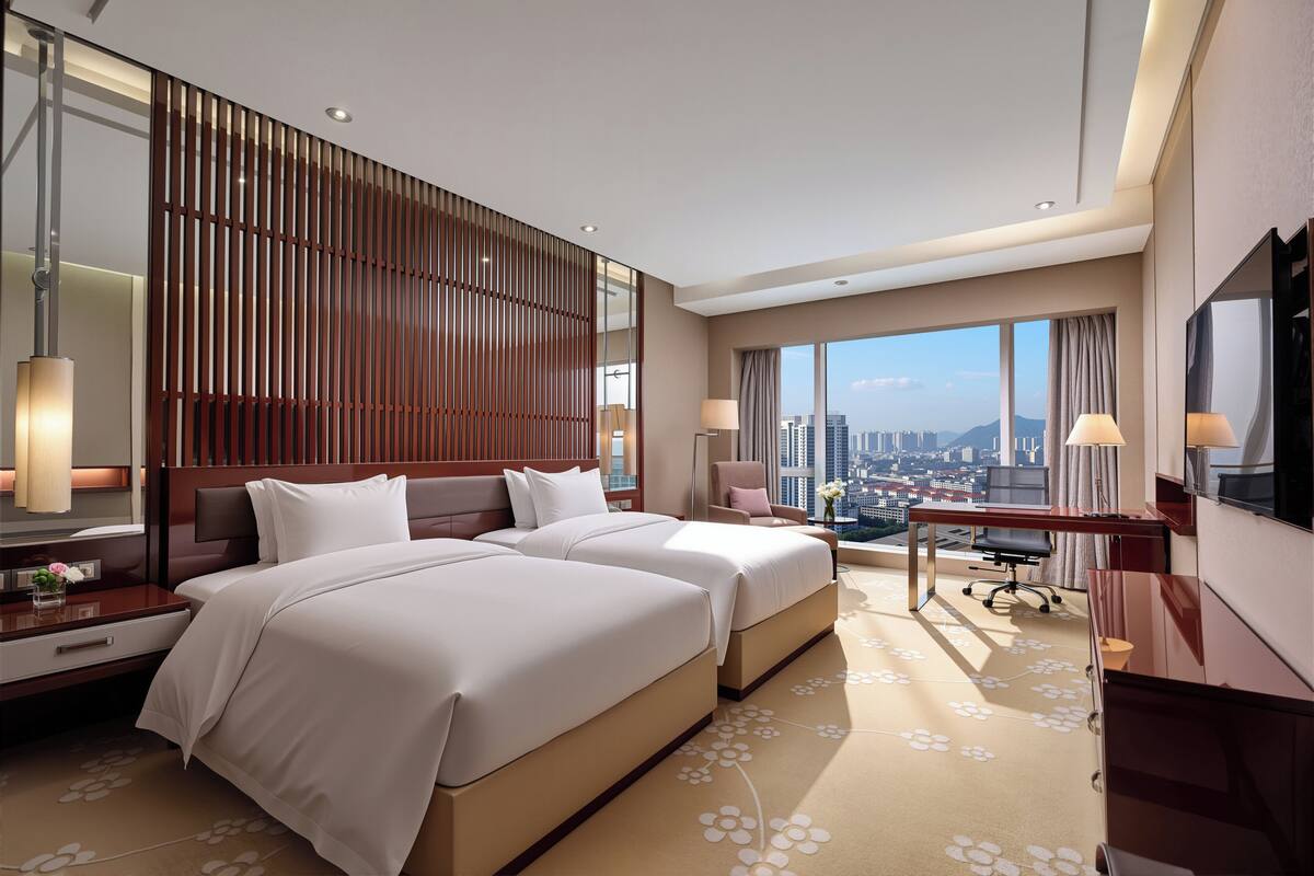 A hotel room with two beds, a desk, a chair, a TV, and a large window with a city view.