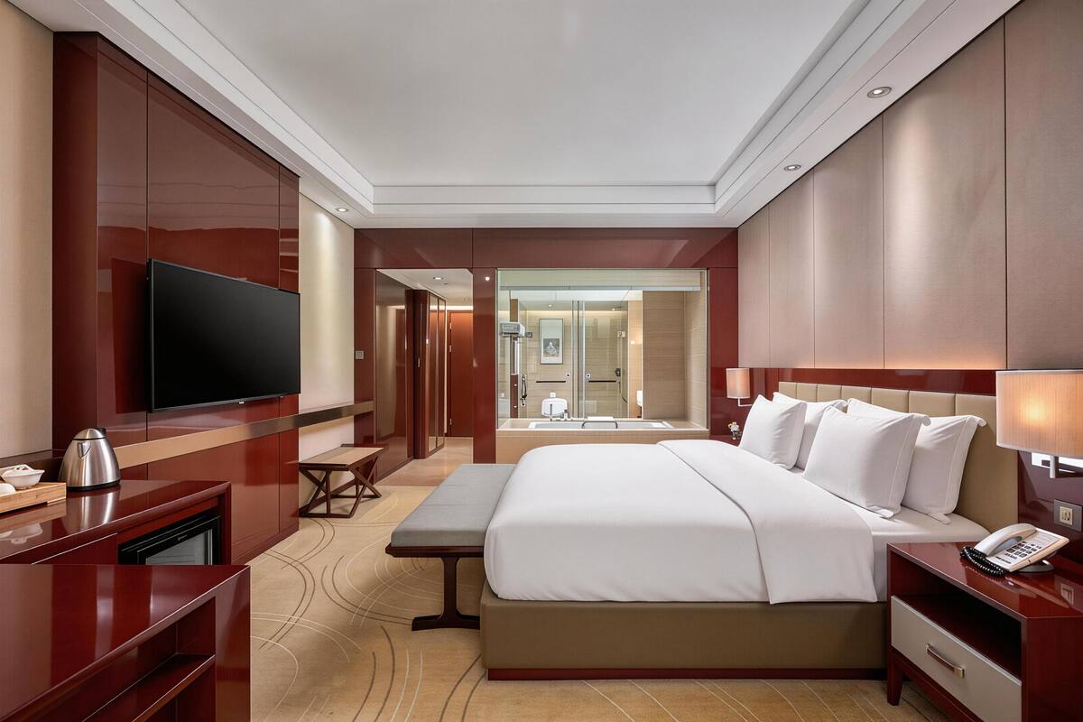 A hotel room with a large bed, a television, a desk with a telephone, and a bathroom visible through an open door.