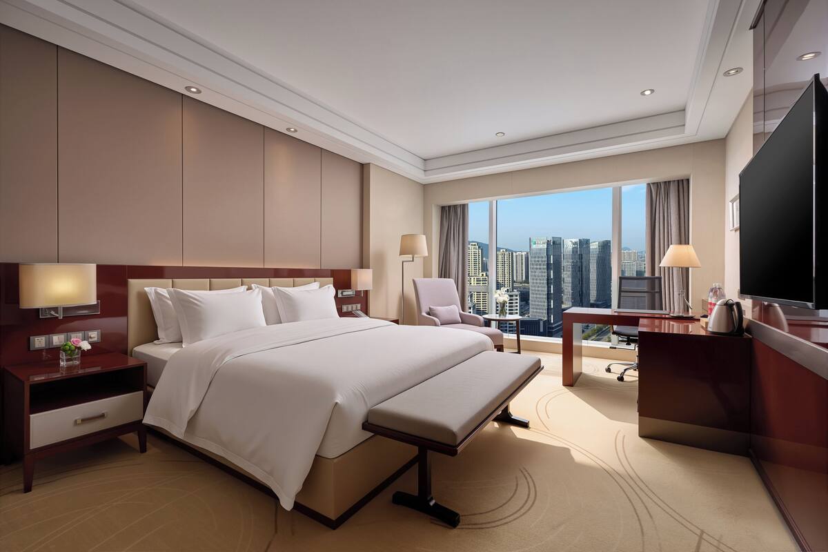 A hotel room with a large bed, bedside tables, a desk, a chair, and a city view.