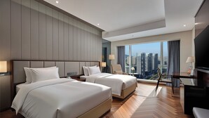 A hotel room with two beds, a TV, a desk, and a large window with a city view.