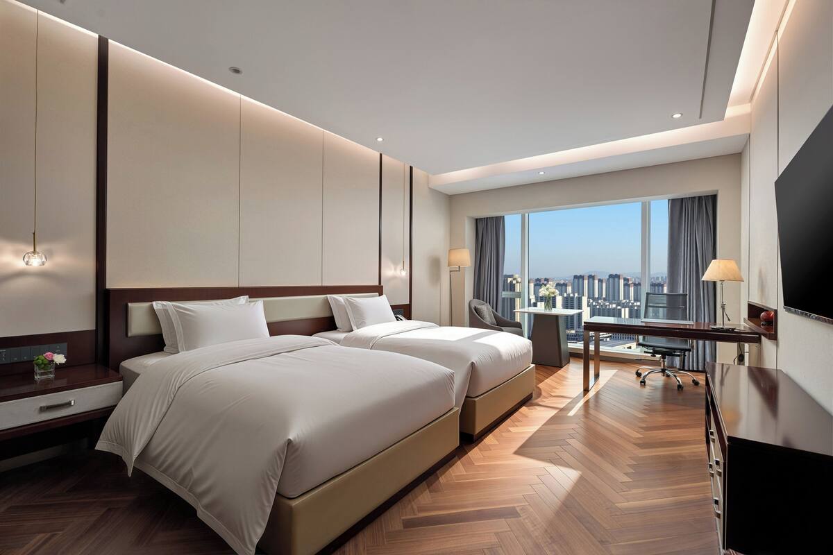 A hotel room with two beds, a desk, a chair, a TV, and a large window with a city view.
