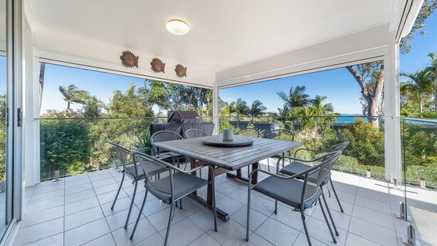 A modern outdoor dining area with a table and chairs, large windows, and a view of palm trees and the ocean. - Oasis 26 (Whitsundays)