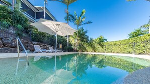 A clear swimming pool with lounge chairs and an umbrella, surrounded by a landscaped area with palm trees and a stone wall. - Oasis 26 (Whitsundays)