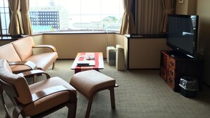 Habitación de hotel con sofá, sillón, reposapiés, una mesita con mandos a distancia, televisor sobre un mueble de madera y una ventana con cortinas.