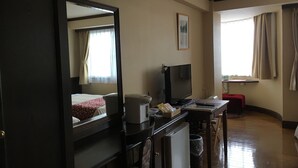 Une chambre d’hôtel comprenant un lit, un bureau avec un ordinateur, une télévision, un petit réfrigérateur, une poubelle et une fenêtre avec des rideaux.