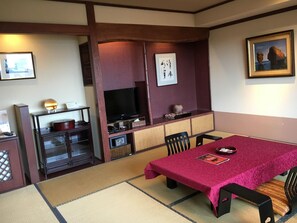 Une pièce de style japonais traditionnel, avec un sol recouvert de tatamis, une petite table recouverte d’une nappe rouge, un téléviseur et un tableau encadré accroché au mur.