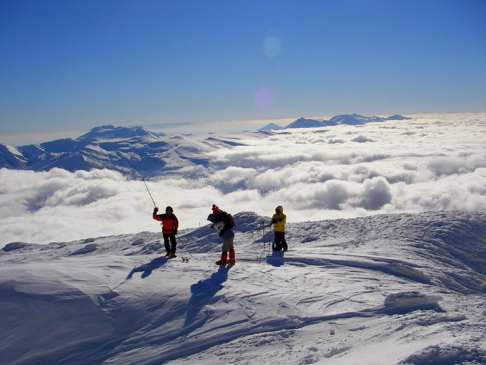 Three people standing on a snowy mountain peak with a clear sky and distant mountains.