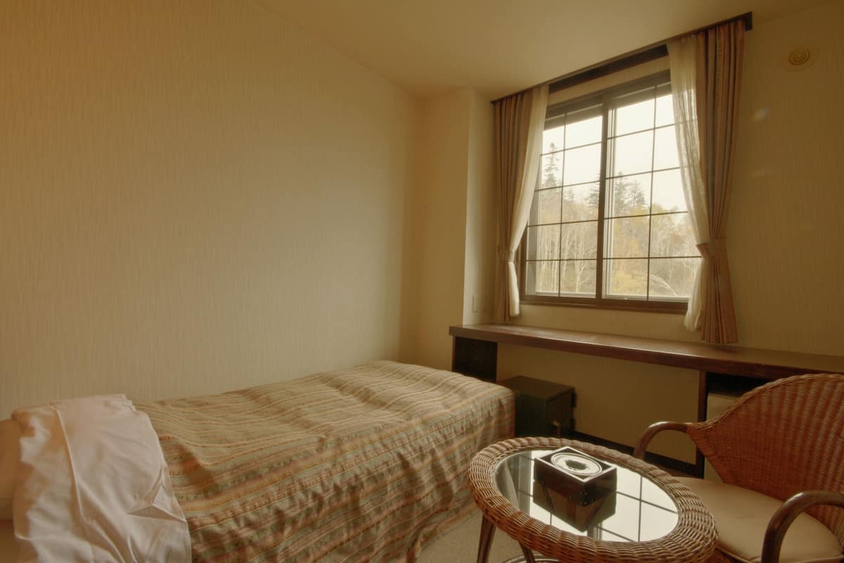 A small, single-room hotel room with a bed, a wicker chair, a small table, and a window with curtains.