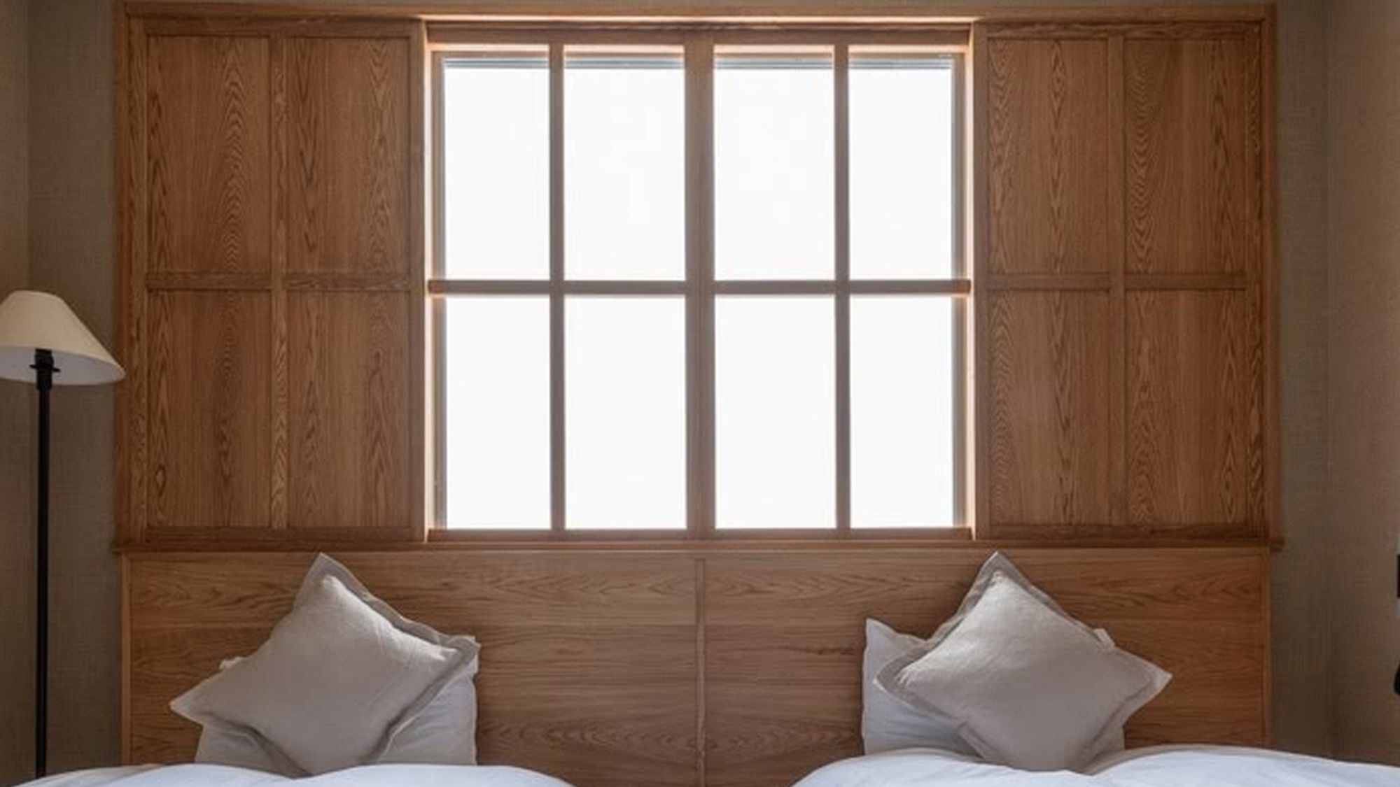 Ein Zimmer mit großem Fenster, Holzwandverkleidung, zwei Betten mit Kissen und einer Stehlampe.