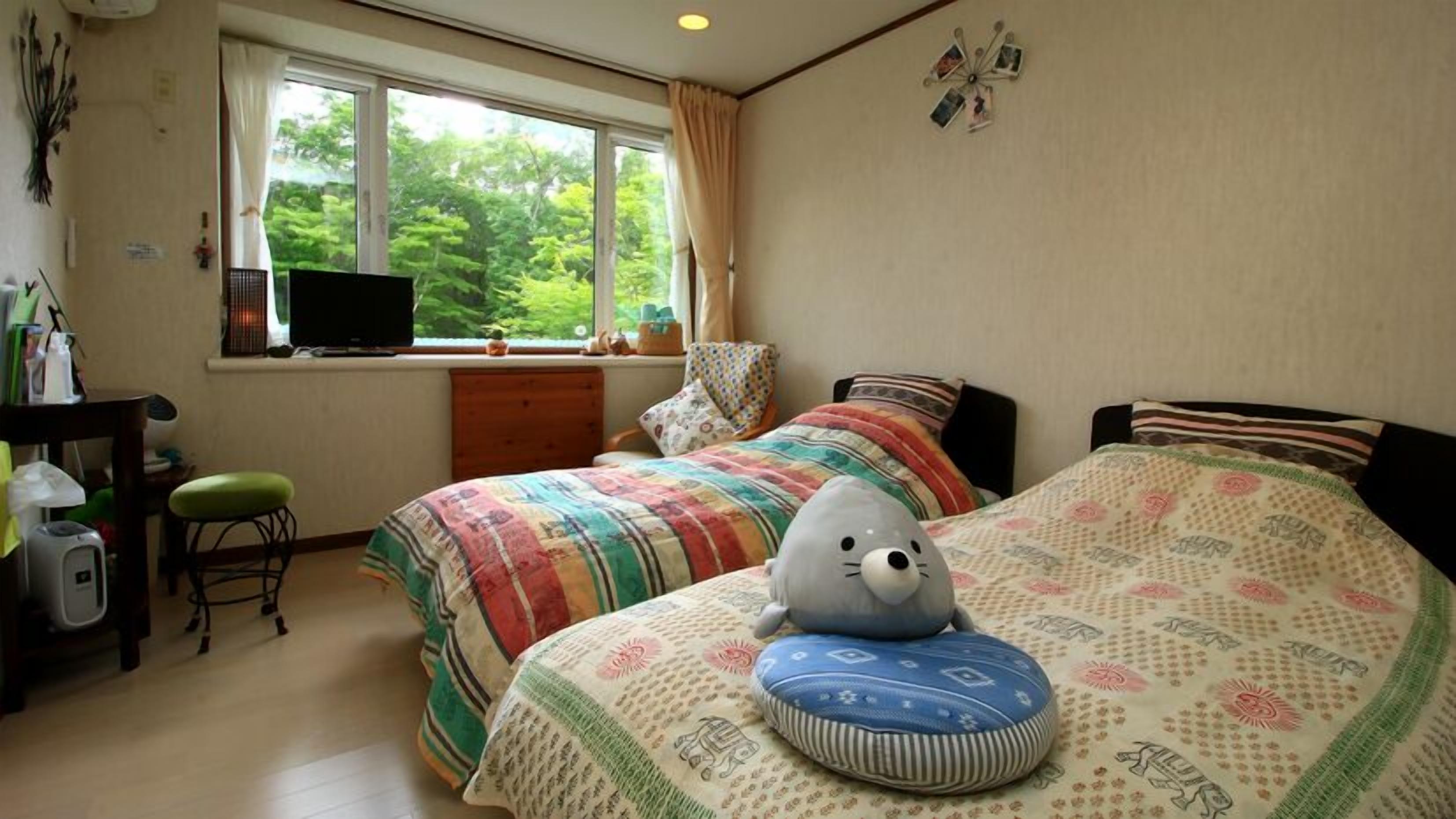 A bedroom with two beds, a desk, a chair, and a window with a view of greenery.