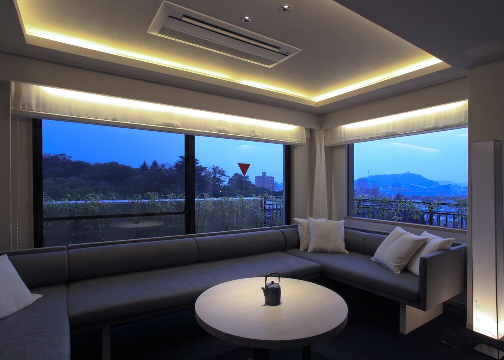 A modern hotel room with a sofa, a round table, and large windows offering a city view.