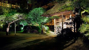 A traditional Japanese-style building with a thatched roof, surrounded by lush greenery and illuminated by ambient lighting. - Dogo Onsen Funaya (Matsuyama)