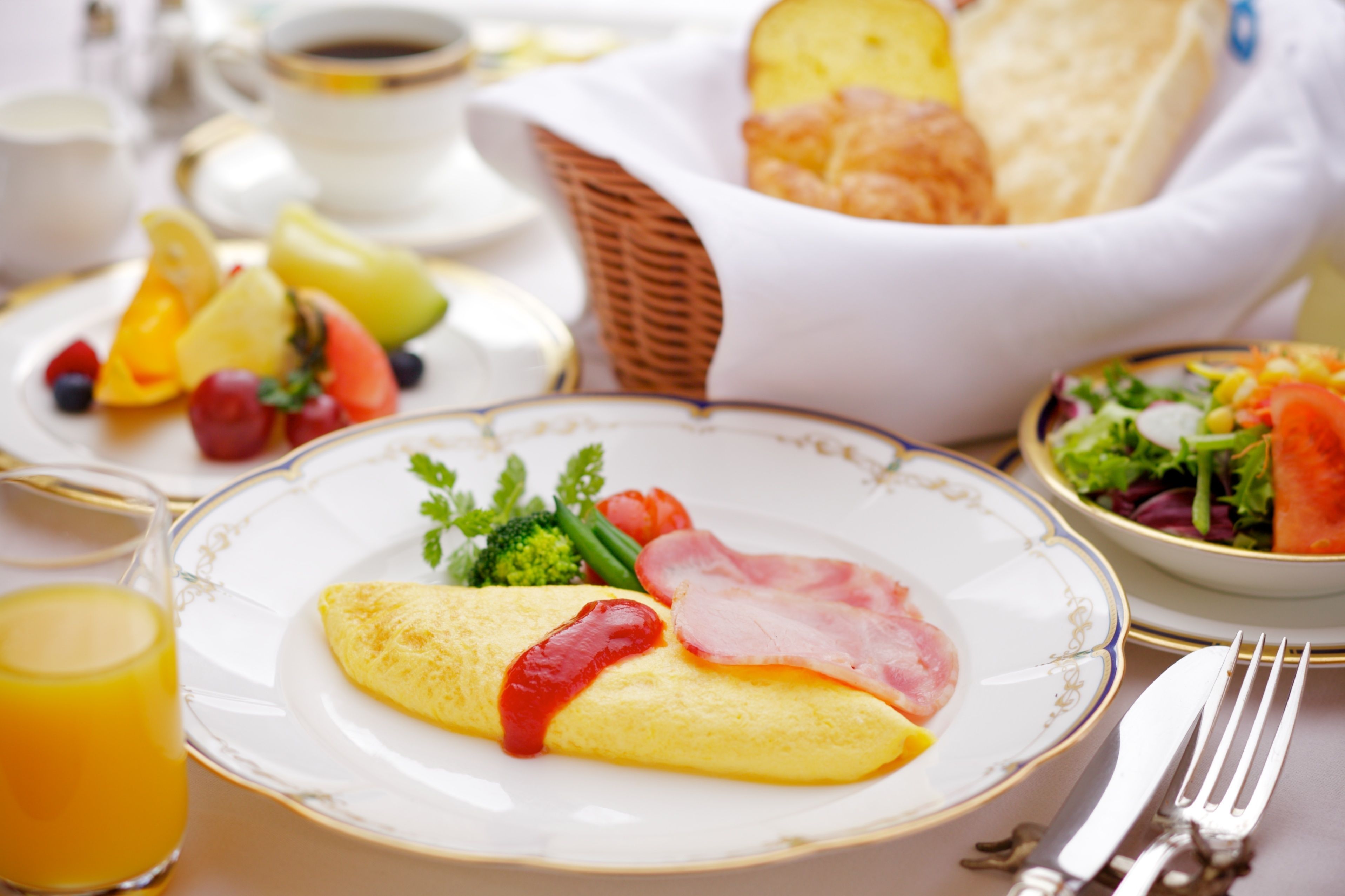 Un desayuno con tortilla, jamón y verduras en un plato, un vaso de zumo de naranja y una cesta de pan.