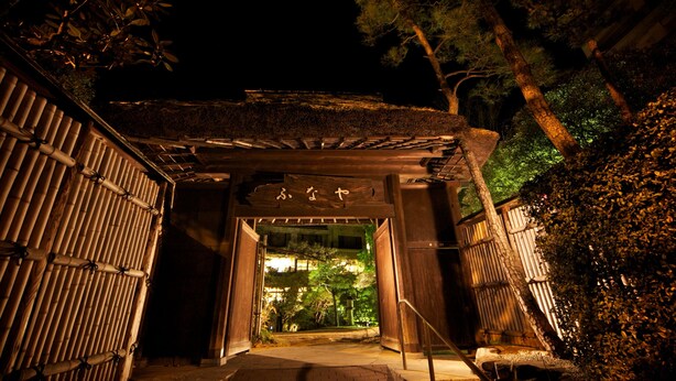 A traditional Japanese gate with a wooden structure and bamboo walls, leading into a well-lit garden area. - Dogo Onsen Funaya (Matsuyama)