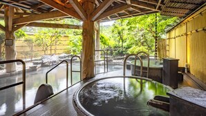 Zona de baños termales con techo de madera, asientos de piedra y vista a un jardín con árboles y una cerca.
