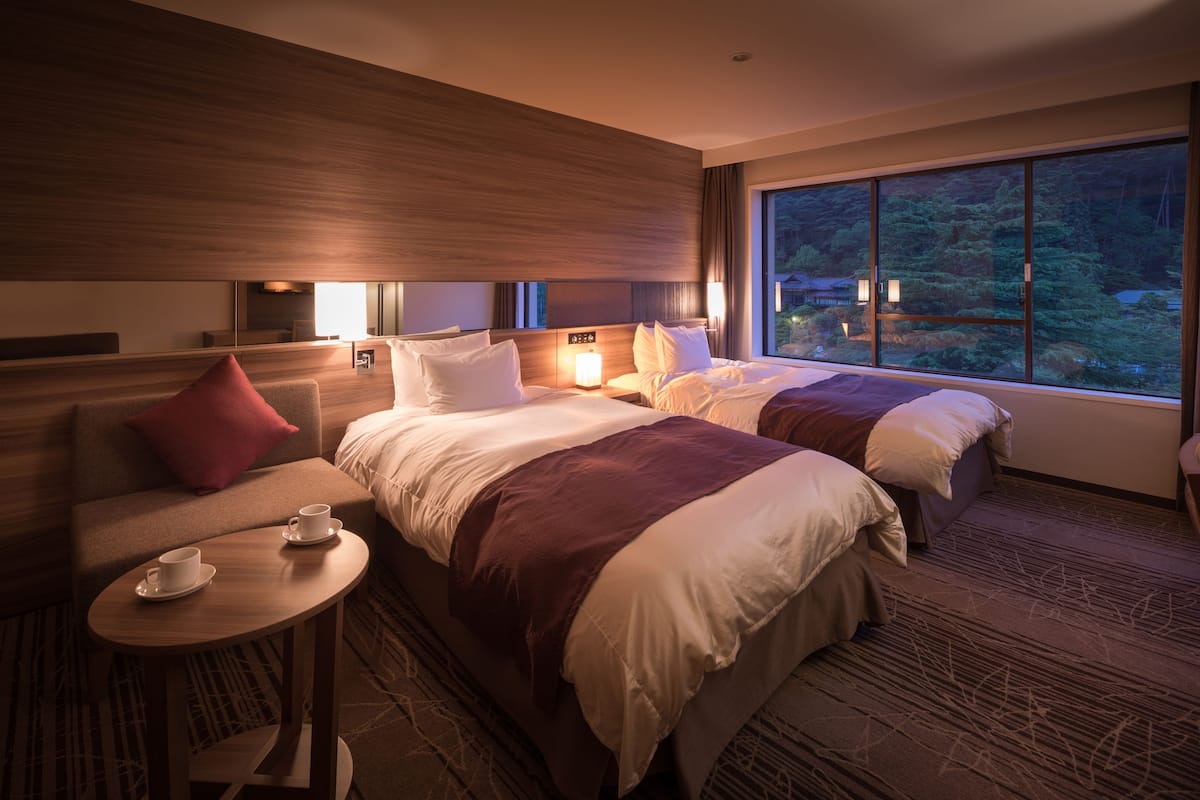 Ett hotellrum med två sängar, en soffa, ett litet bord med koppar och ett stort fönster med utsikt över träd.