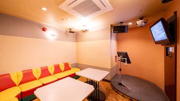 A small, cozy room with a red and yellow booth seating area, two white tables, a small stage with a microphone stand, and a flat-screen TV mounted on the wall.
