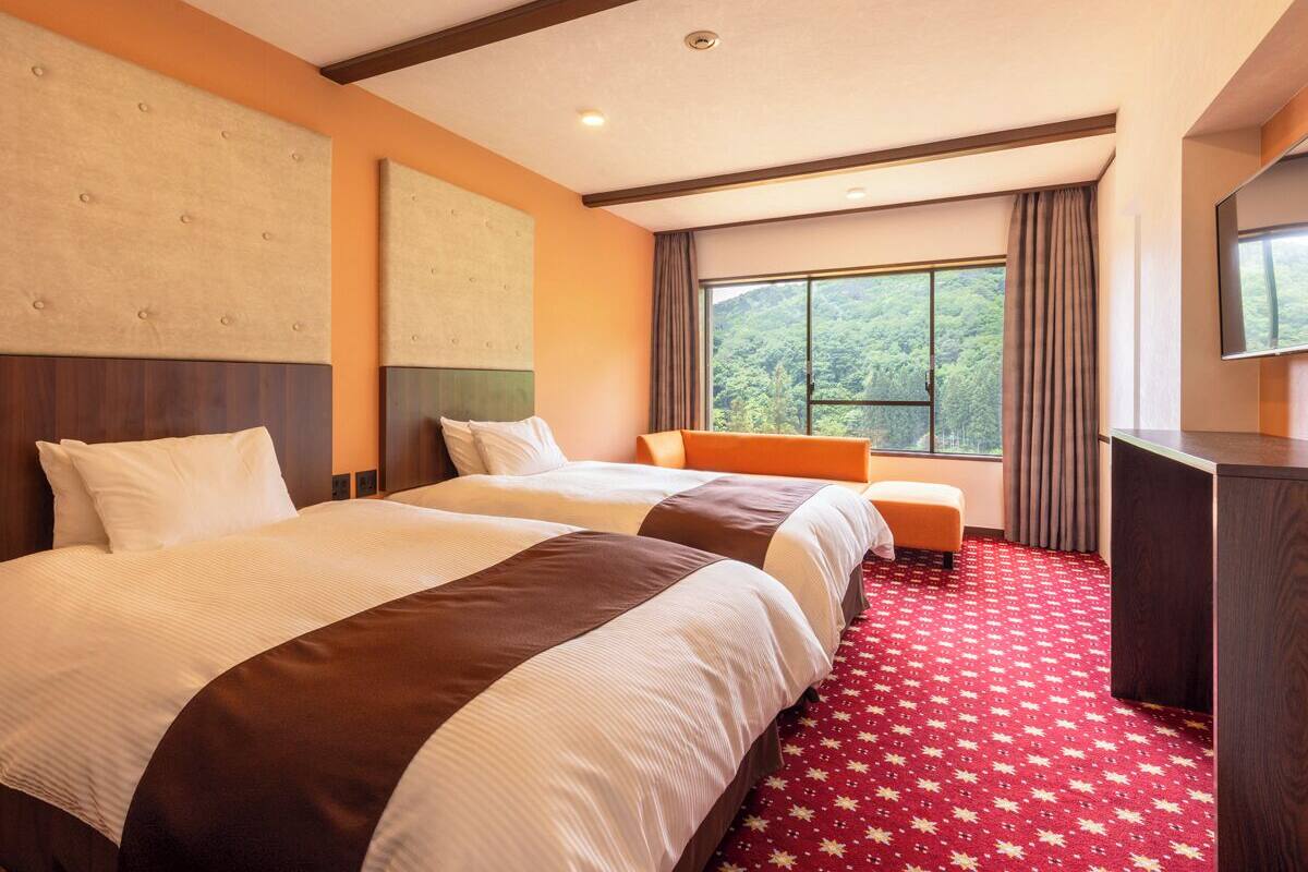 Ett hotellrum med två sängar, ett skrivbord och ett stort fönster med utsikt över grönområden.