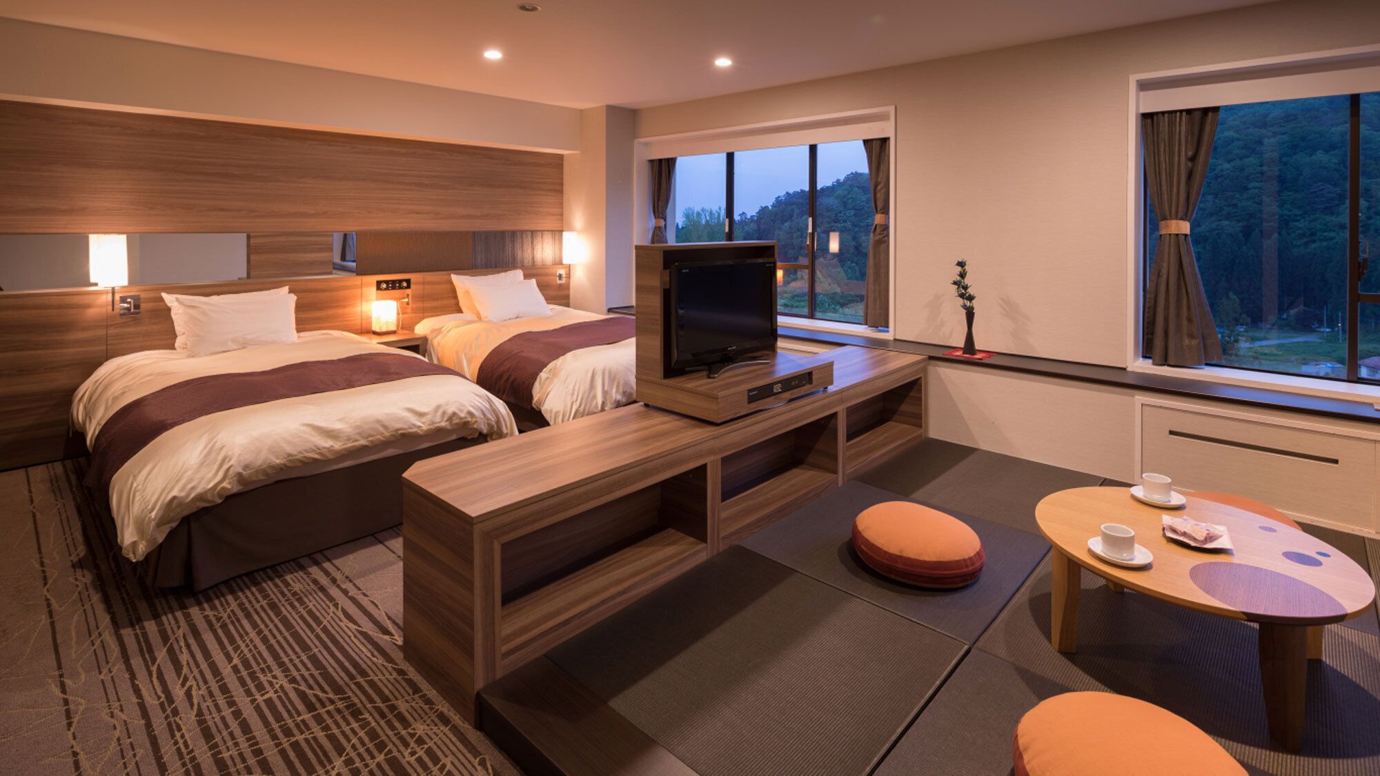 A hotel room with two beds, a TV, a small table, and a window with a view of trees.