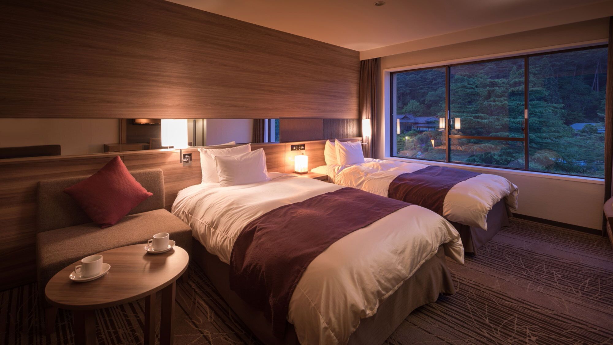 A hotel room with two beds, a sofa, a small table with a cup, and a large window with a forest view.
