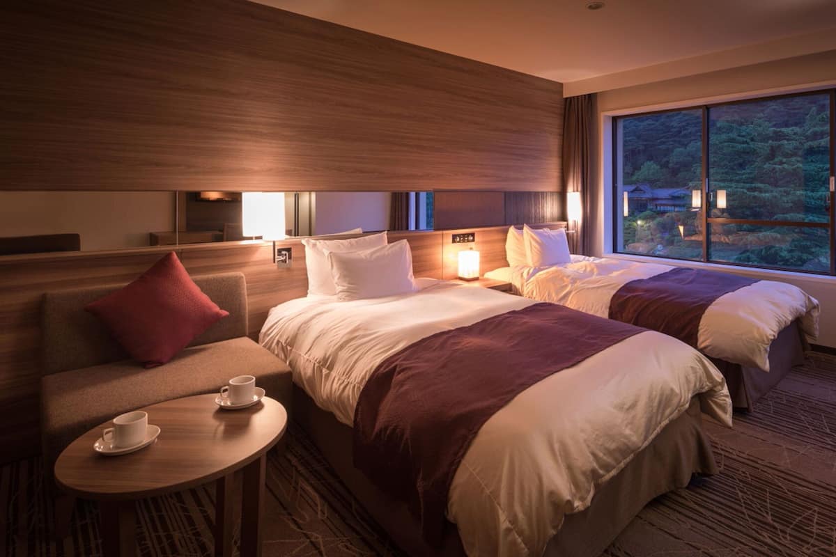 A hotel room with two beds, a sofa, a small table with a cup, and a large window with a forest view.