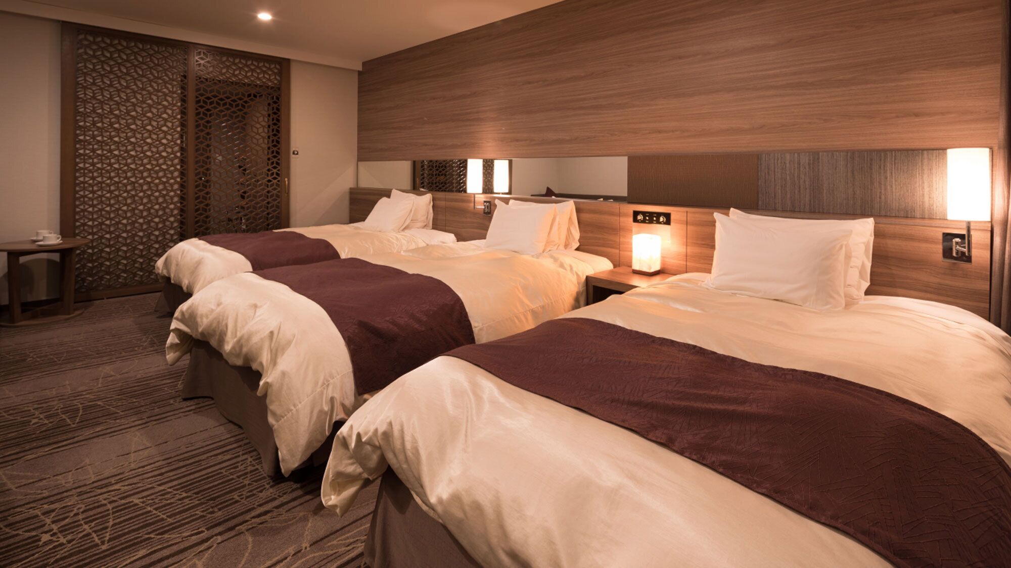 A hotel room with three beds, a wooden headboard, a nightstand with a lamp, and a decorative wall panel.
