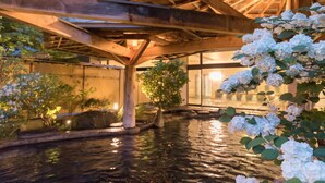 Un jardín interior tranquilo con fuente de agua, vigas de madera y abundante vegetación.