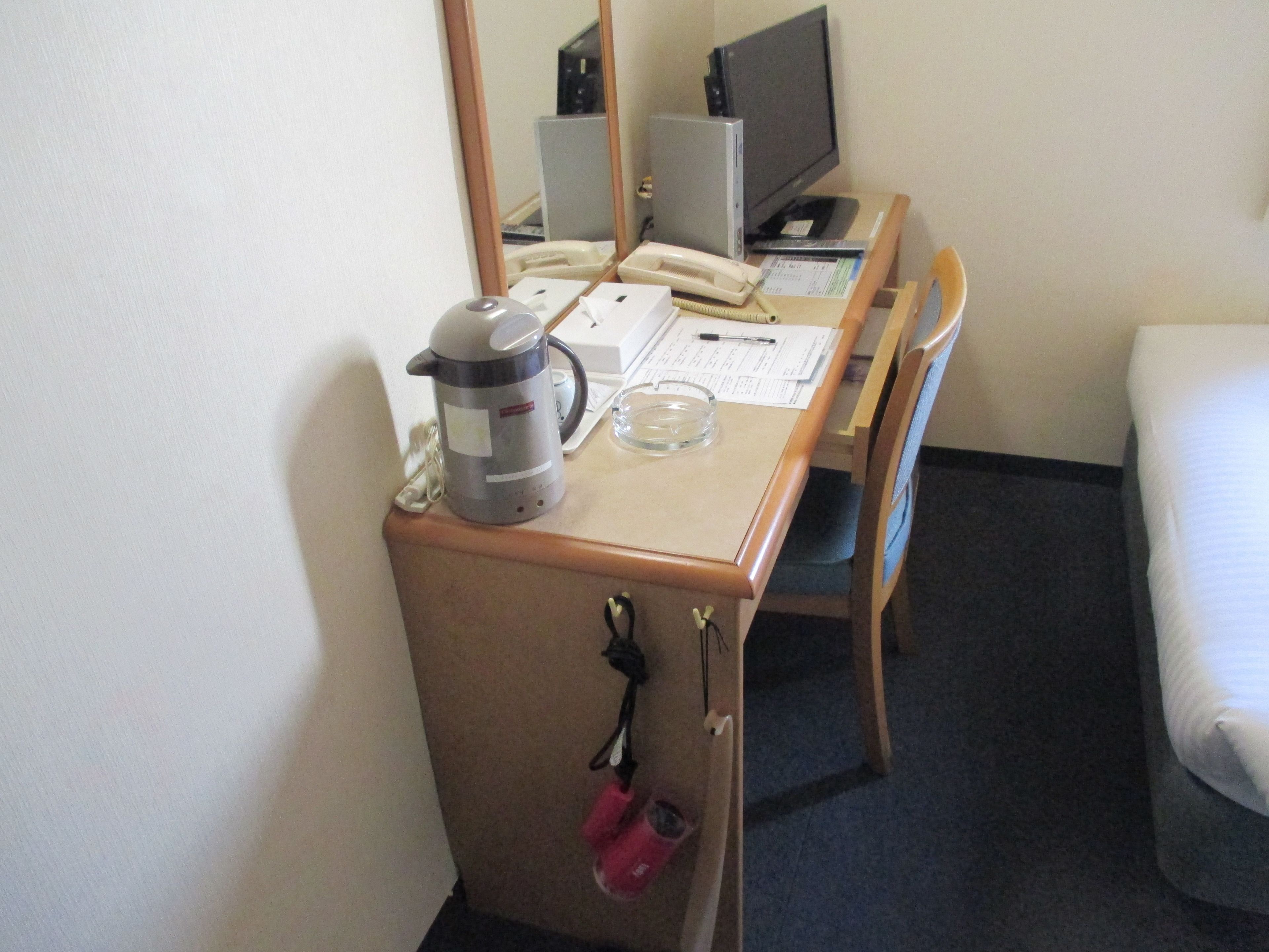 A hotel room with a wooden desk, a computer, a telephone, a kettle, and a bed.