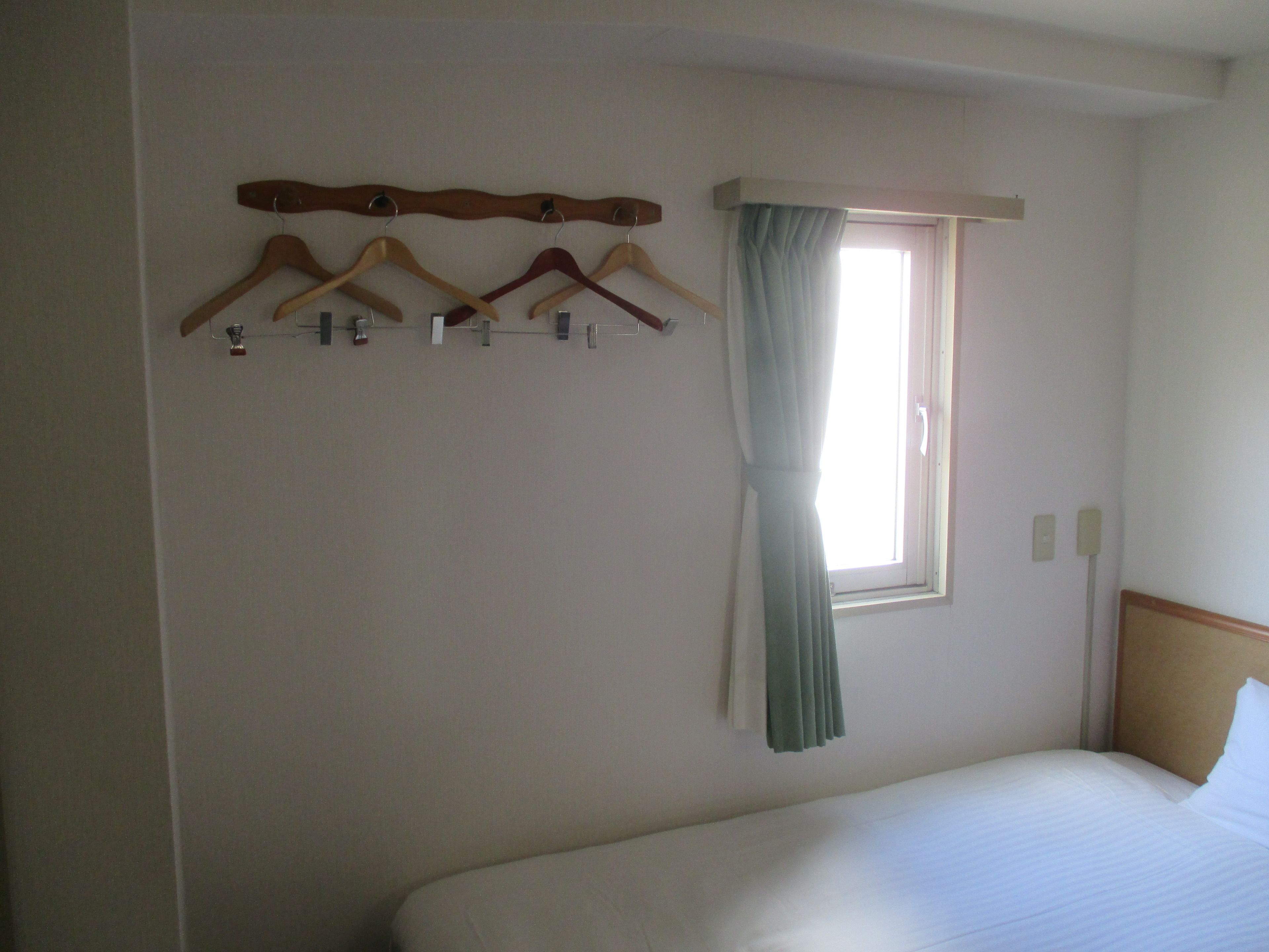 Una habitación de hotel con cama, ventana con cortinas y un perchero con perchas.
