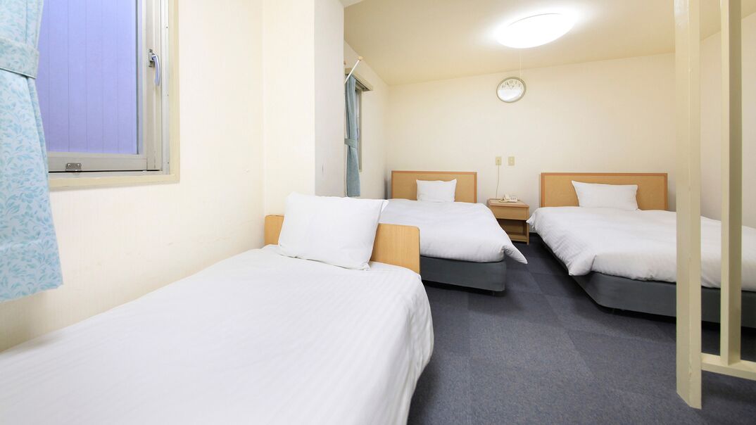 A hotel room with three single beds, a bedside table with a lamp, a window with blinds, and a clock on the wall.