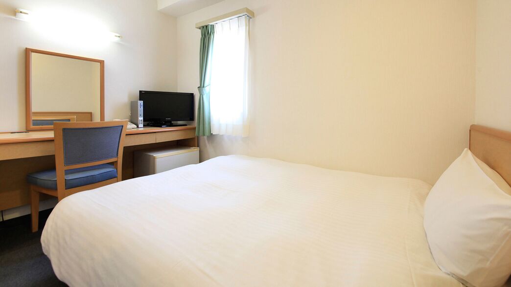 A hotel room with a bed, a desk with a computer, a chair, and a window with curtains.