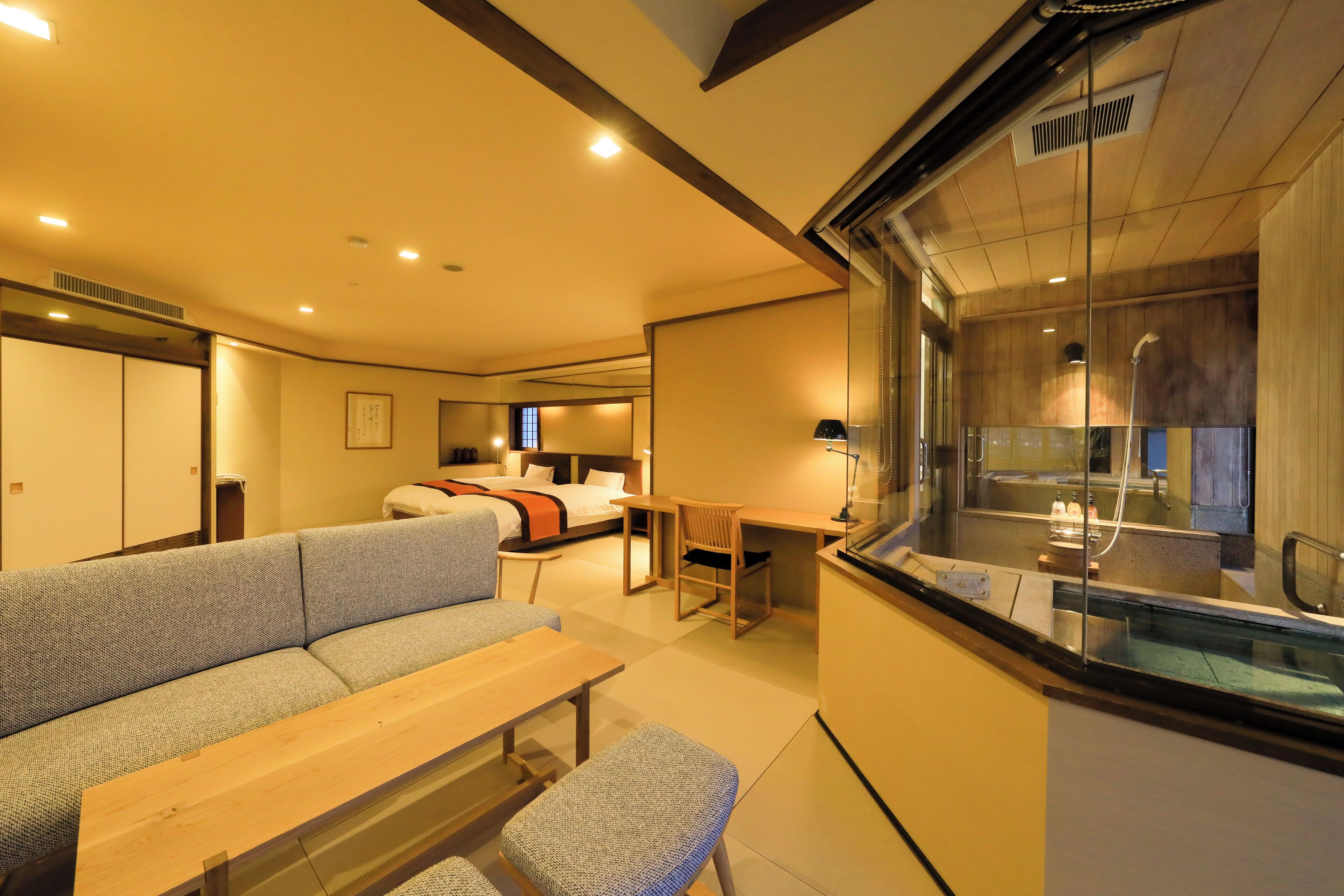 A modern hotel room with a sofa, a wooden table, a bench, a desk, and a glass partition separating the living area from the bathroom.