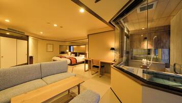 A modern hotel room with a sofa, a wooden table, a bench, a desk, and a glass partition separating the living area from the bathroom.