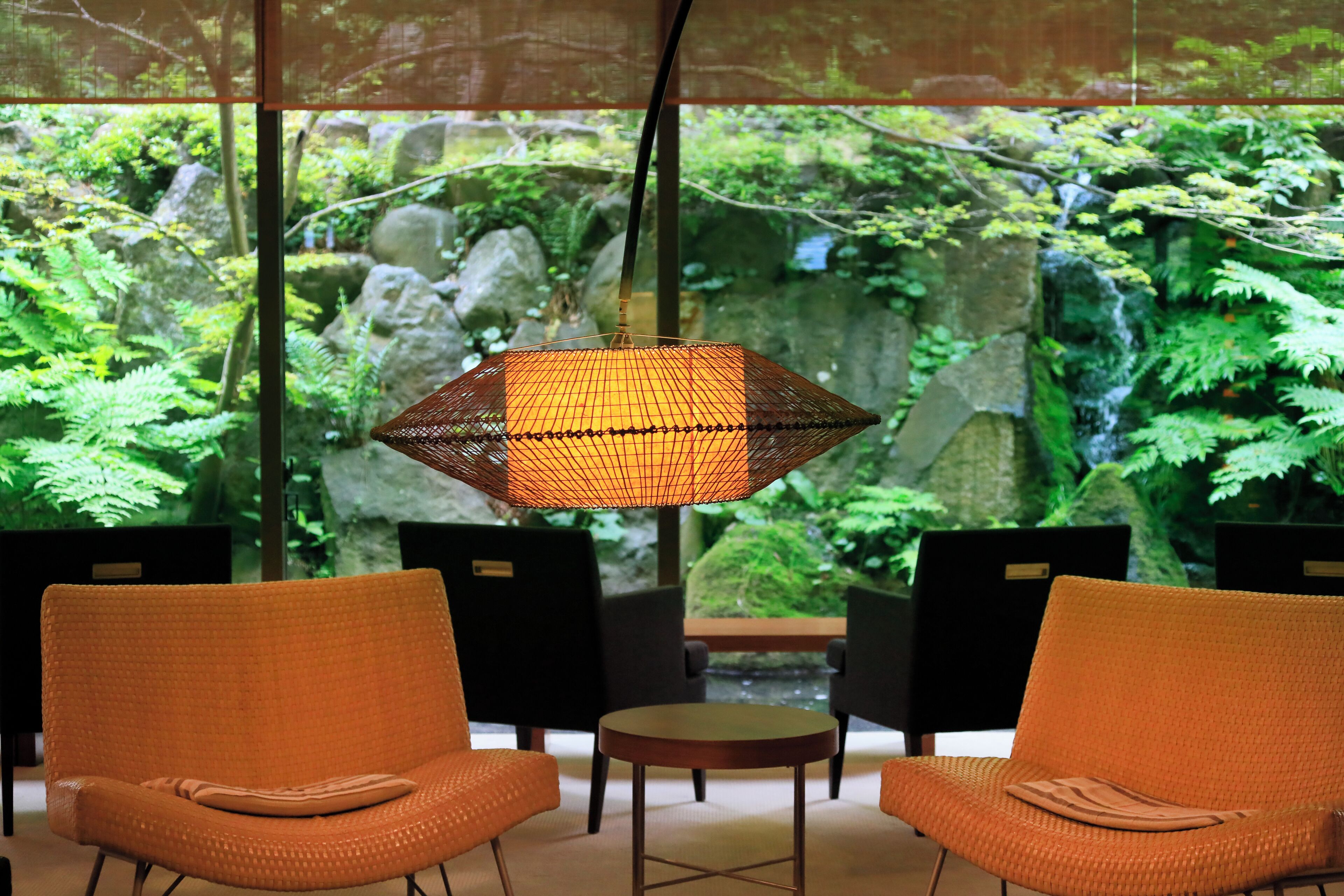 Una habitación con dos sillones de mimbre naranja, una mesita redonda pequeña y una lámpara colgante. Un ventanal muestra un jardín verde exuberante con rocas y helechos.