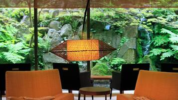 Una habitación con dos sillones de mimbre naranja, una mesita redonda pequeña y una lámpara colgante. Un ventanal muestra un jardín verde exuberante con rocas y helechos.