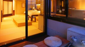 Una habitación de hotel moderna con una puerta corrediza de vidrio que da a un balcón, una cama, una pequeña zona de estar y un baño visible a través del vidrio.