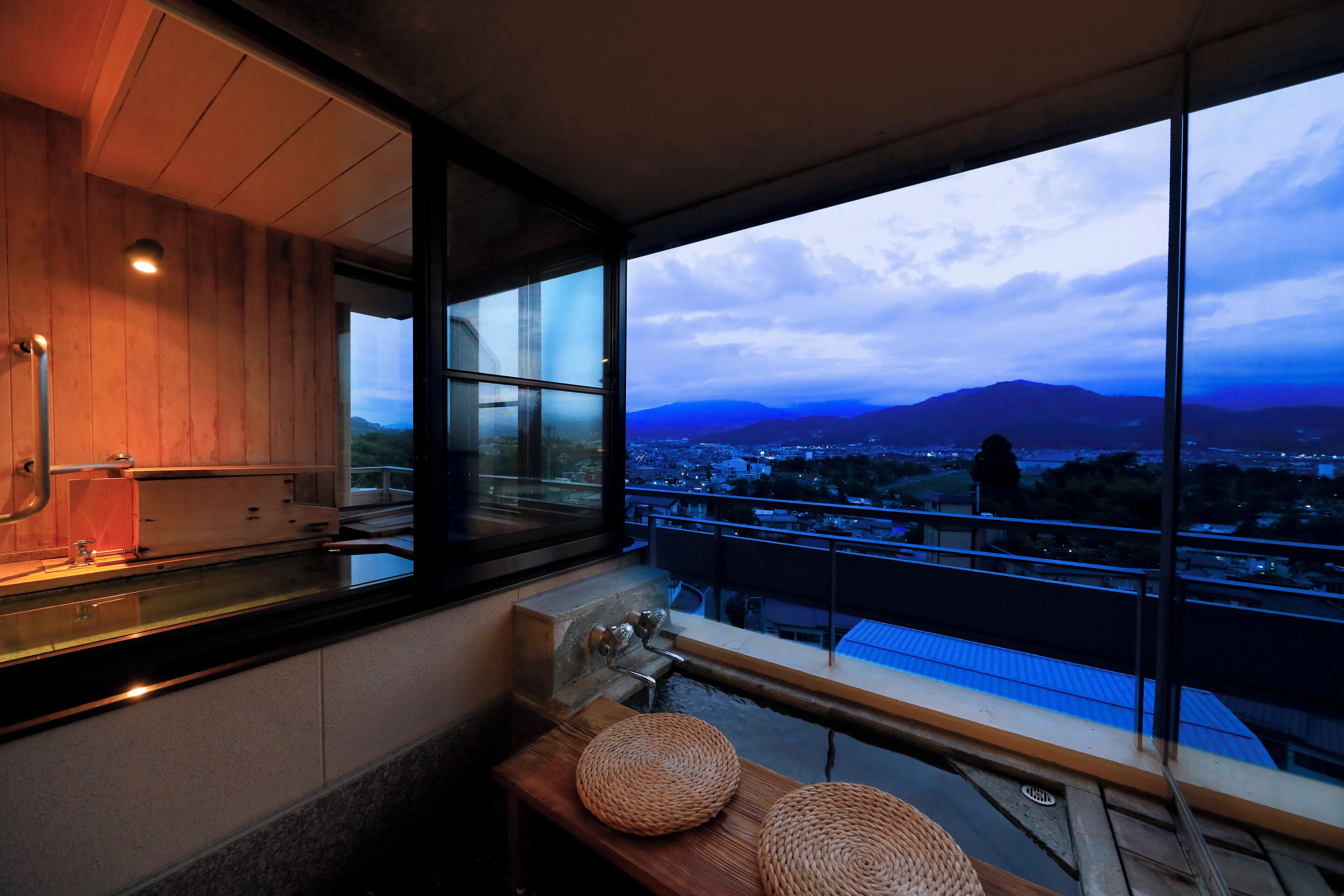 Un baño moderno con un tocador de madera, un lavamanos y un amplio ventanal con vistas a un paisaje urbano y montañas.