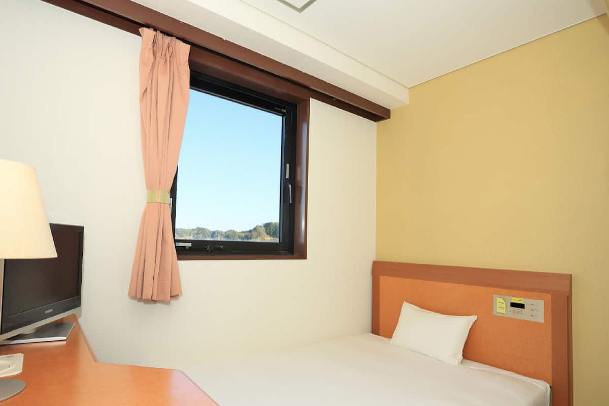 A hotel room with a single bed, a desk with a lamp, a window with a view of the landscape, and a thermostat on the wall.