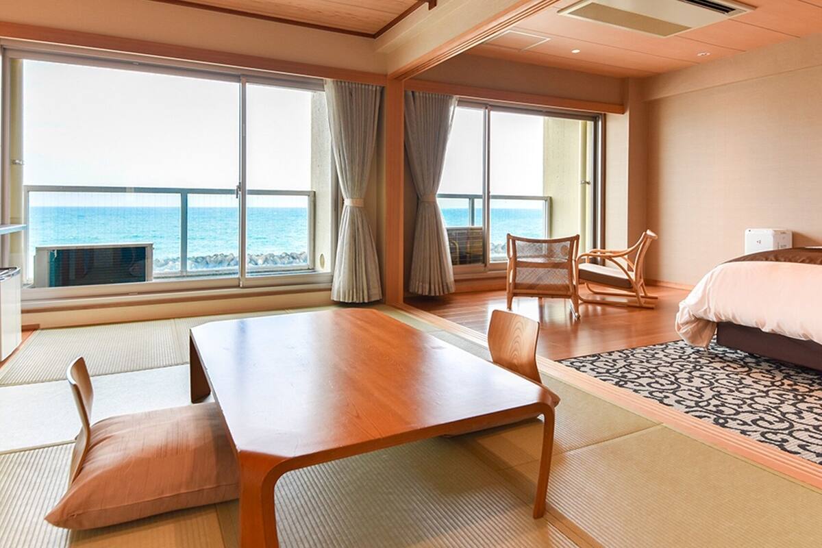 A hotel room with a wooden table, two chairs, a bed, and large windows with ocean views.