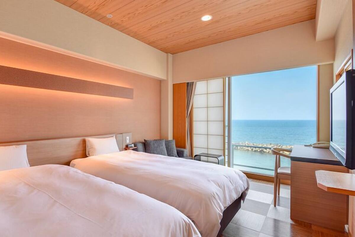 A hotel room with two beds, a desk, and a view of the sea.