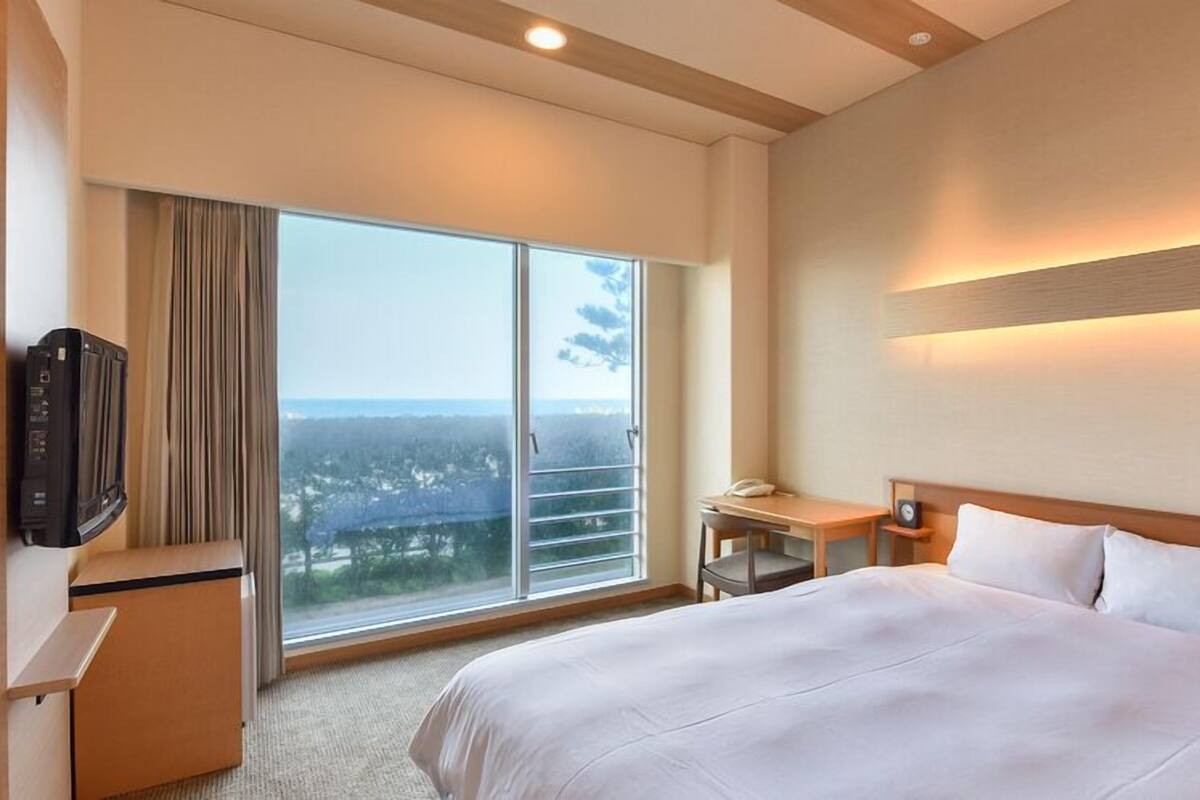 A hotel room with a large bed, a desk, a chair, a TV, and a view of the city through a large window.