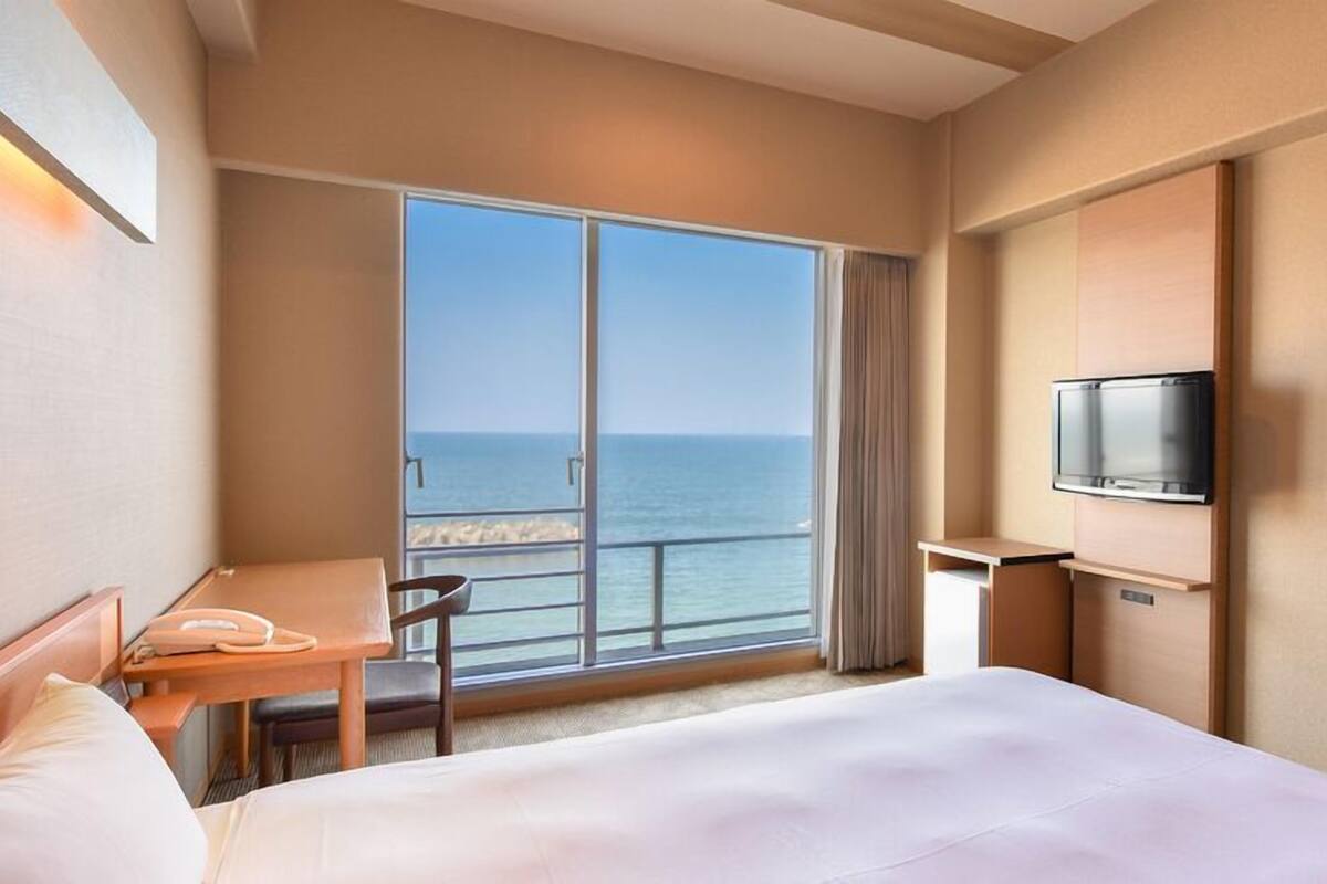A hotel room with a bed, a desk, a chair, a television, and a large window with a view of the ocean.