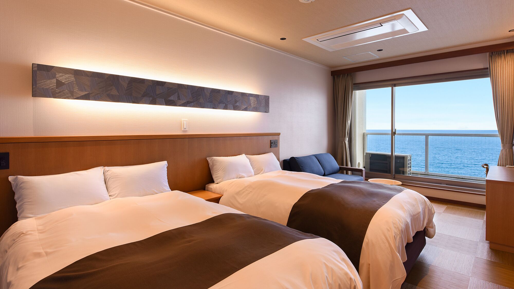 A hotel room with two beds, a large window with a view of the sea, a ceiling light, and a wall-mounted artwork.