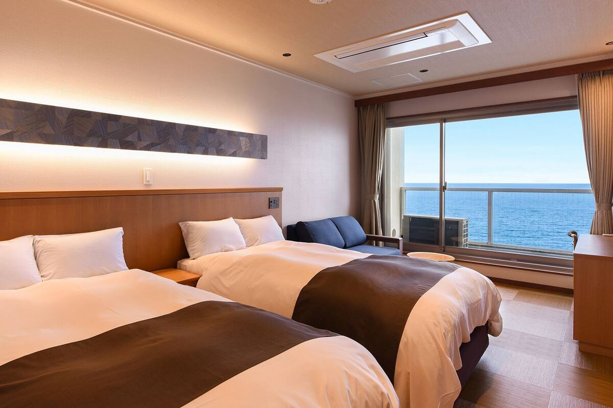 A hotel room with two beds, a large window with a view of the sea, a ceiling light, and a wall-mounted artwork.