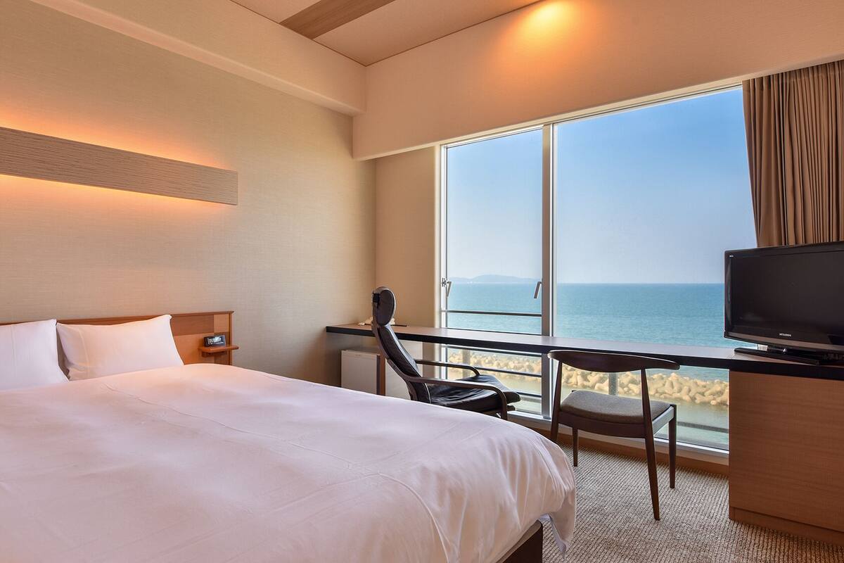 A hotel room with a bed, a desk with a chair, a television, and a large window with a view of the sea.