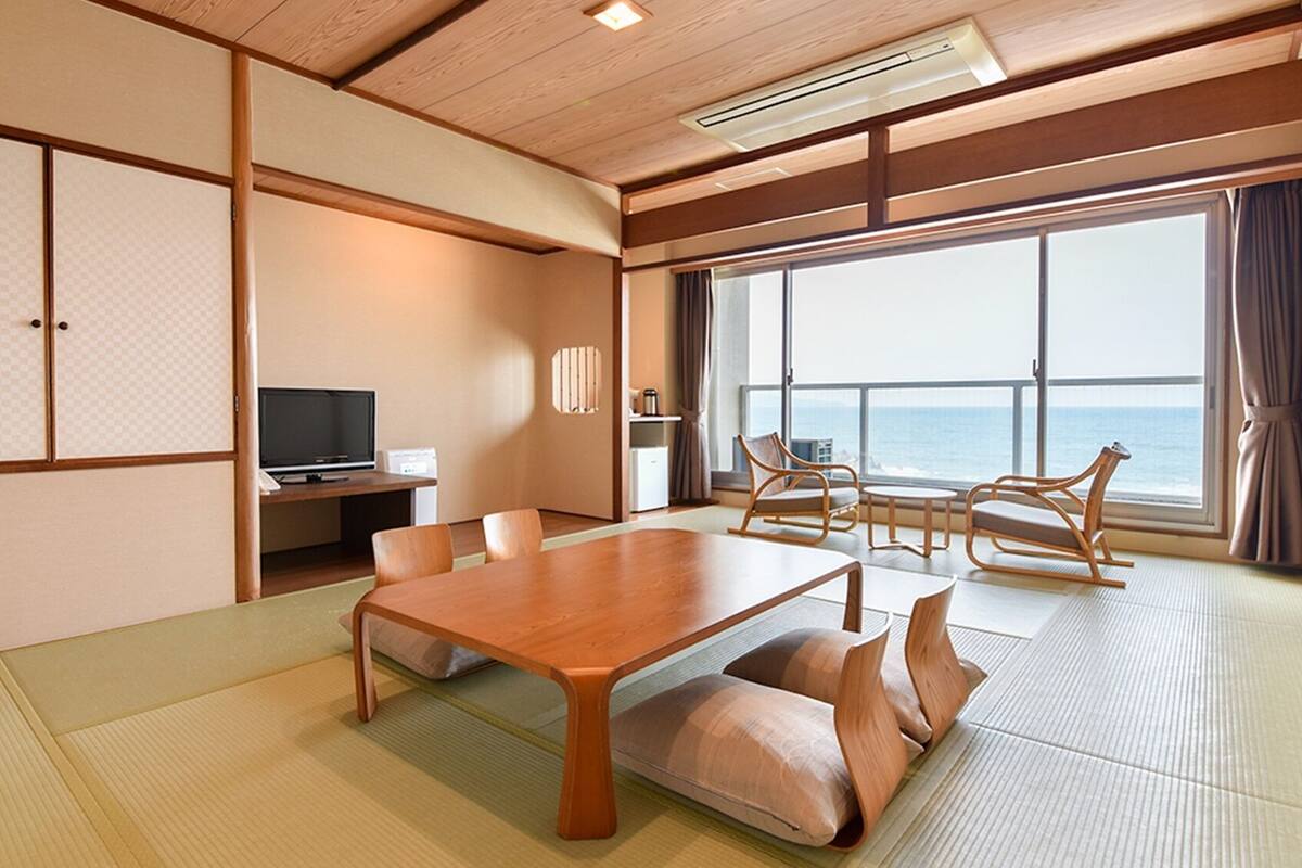 A traditional Japanese-style room with tatami flooring, a wooden table, chairs, a television, and a view of the ocean.